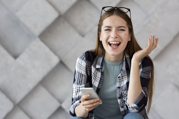 Young woman holding smartphone, laughing and wearing glasses on head, illustrating Gen Z handing out compliments creatively.