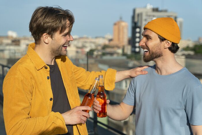 Two men smiling and clinking bottles outdoors, sharing Gen Z compliments and positive vibes on a sunny day.