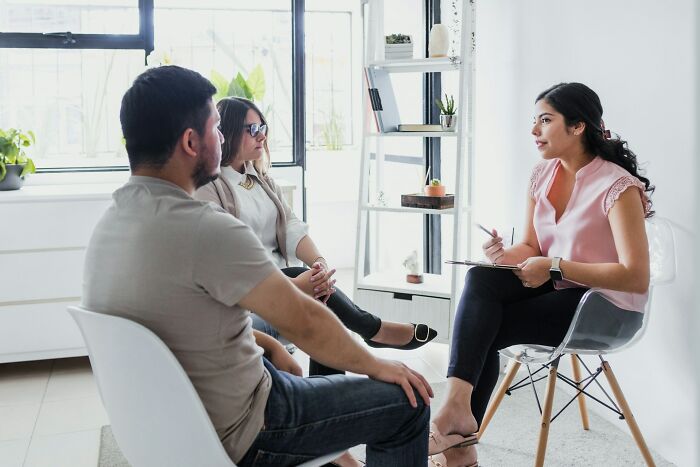 Couple discussing relationship issues with a counselor in a bright room, addressing partner's shenanigans and conflicts.