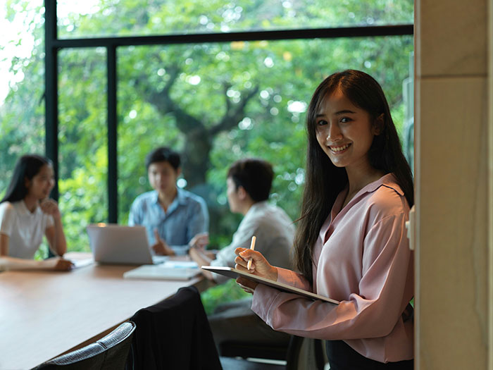 Young woman holding a tablet, looking determined while others work in a bright room with large windows.