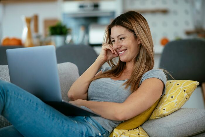 Woman smiling while using a laptop on a couch, representing small daily wins from feminists in a relaxed home setting.