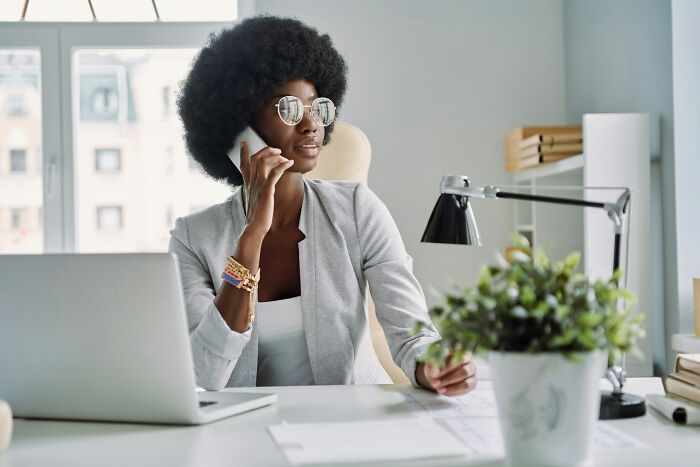 Woman with afro hairstyle wearing sunglasses and blazer, talking on phone at a desk, representing feminist daily wins.