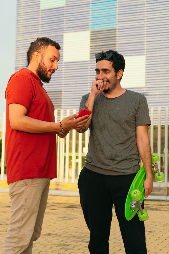 Two men sharing a funny moment outdoors, one holding a skateboard, illustrating things normal to men but bizarre to women.
