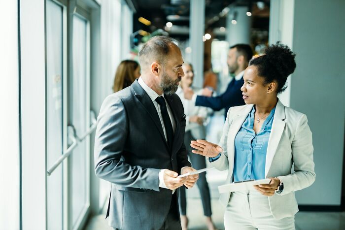 Businesswoman discussing small daily wins from feminists with male colleague in a modern office hallway during workday.