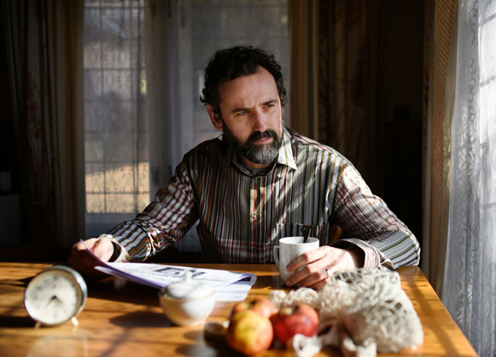 A man with a beard sitting at a table, holding a cup, reflecting on woman deciding to leave partner homeless.