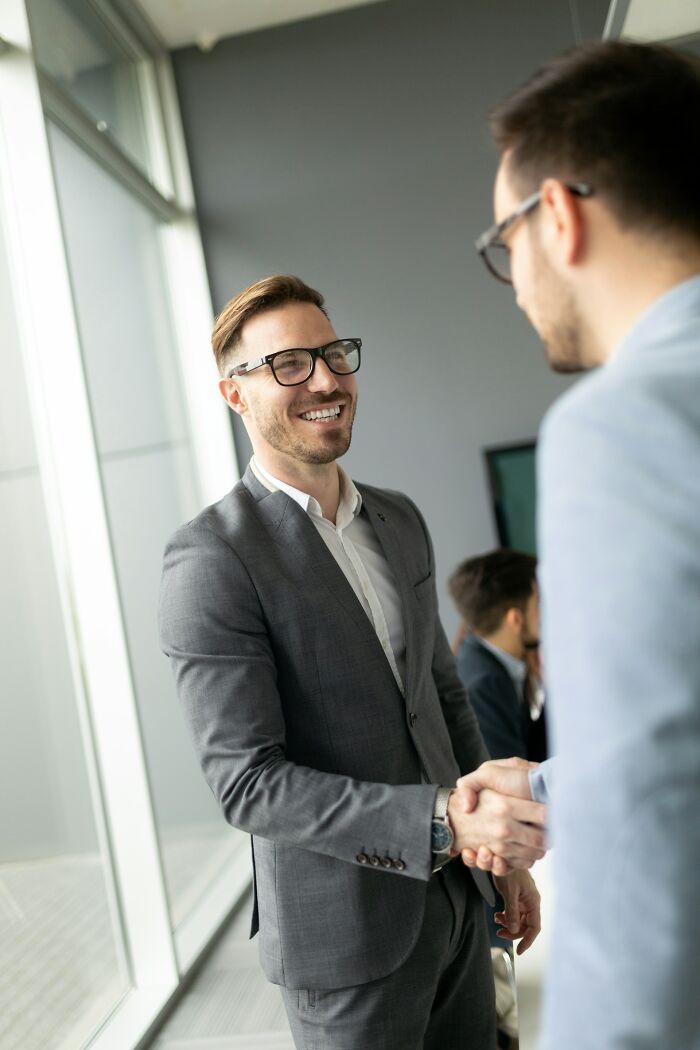 Two men in business attire shaking hands, smiling, sharing hard-to-believe stories about themselves in an office setting.