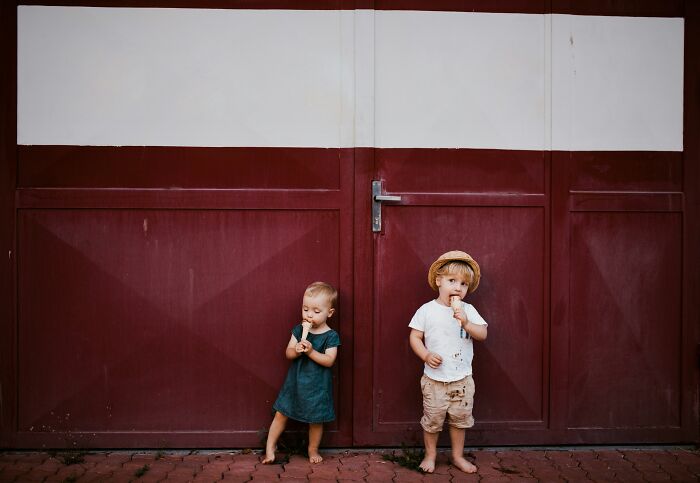 Two young children eating ice cream while standing barefoot against a large maroon door, symbolizing stuck relationships and longing.