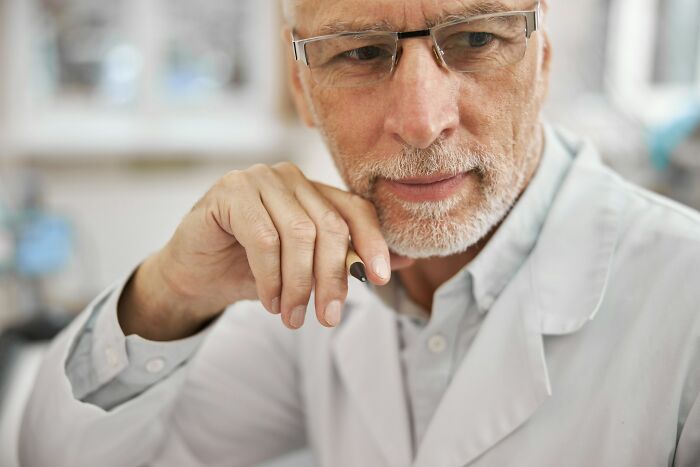 Older doctor wearing glasses and a white coat, looking thoughtful while holding a pen in a medical office.