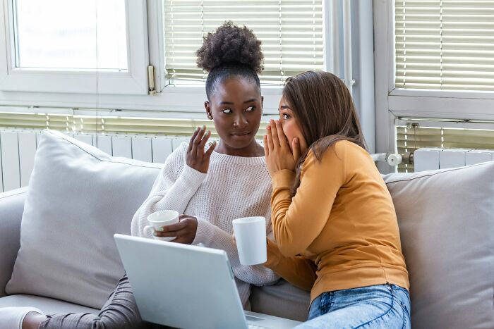 Two women seated on a couch sharing a secret while drinking coffee, illustrating toxic femininity behaviors concept. - 21
