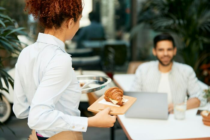 Waitress serving croissant to man working on laptop in cafe, highlighting small daily wins from feminists.