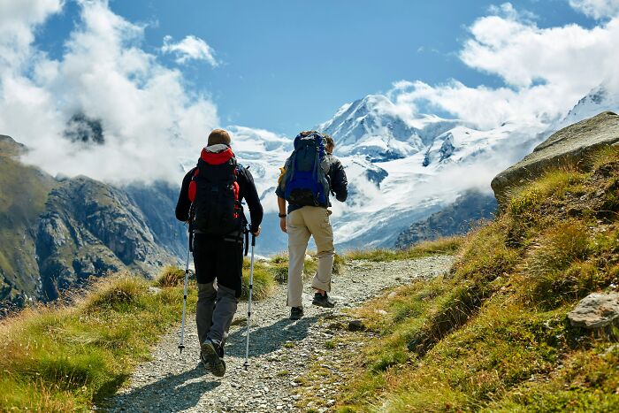 Two hikers walking a mountainous trail under a blue sky, unrelated to people who deal with dead bodies or weird discoveries.