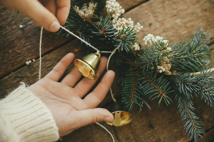 Hand holding a small golden bell near a pine branch decorated with white berries, for student names article.