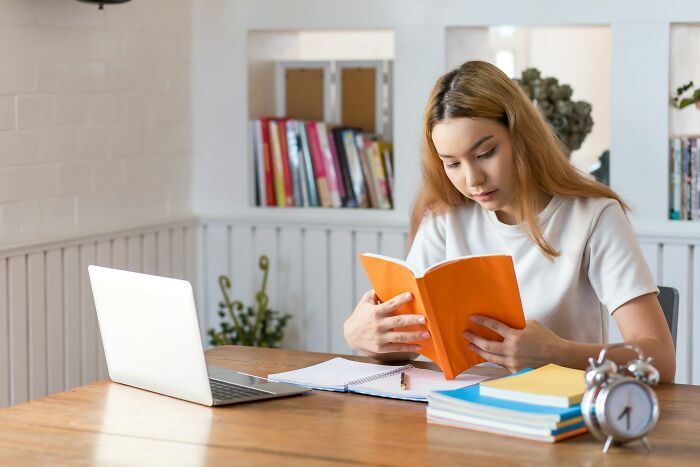 Young woman reading a book at a wooden table with laptop and notebooks, symbolizing stories of people who ran away from home. - 11