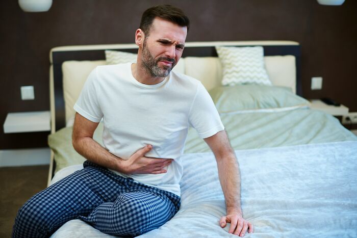 Man in pajamas sitting on bed, holding stomach in pain, showing side effects eating watermelon like discomfort and cramps. - 6