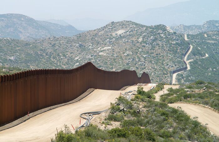 Border wall extending through mountainous terrain, illustrating bizarre facts about countries shared by 80 people.