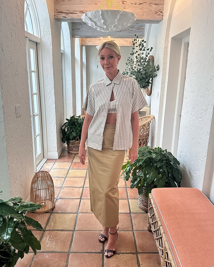 Gwyneth Paltrow standing in a hallway, wearing a striped shirt and beige skirt, before a cooking video on trash handling. - 1