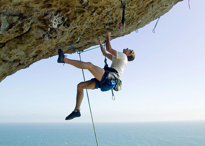 Man rock climbing on a steep cliff overlooking the ocean, illustrating risks linked to habits and decisions that reduce life quality.