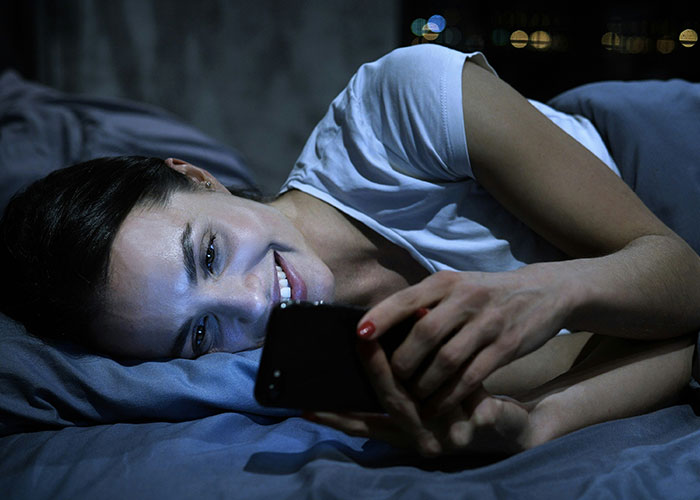 Woman lying in bed at night smiling while using a smartphone, illustrating habits impacting life quality and decisions.
