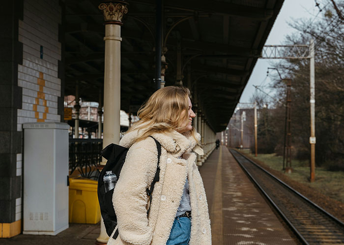 Young woman at a train station platform, wearing a coat and backpack, reflecting on habits that reduce life quality.