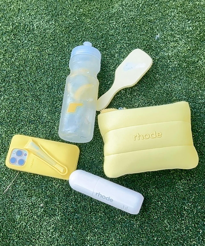 Yellow Rhode beauty pouch, water bottle, phone case, and white cylindrical bedazzled tampon accessory on green grass.