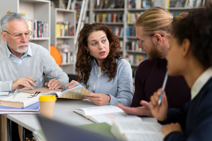 Group of students and adults discussing and studying together around a table in a school hallway or lane setting.