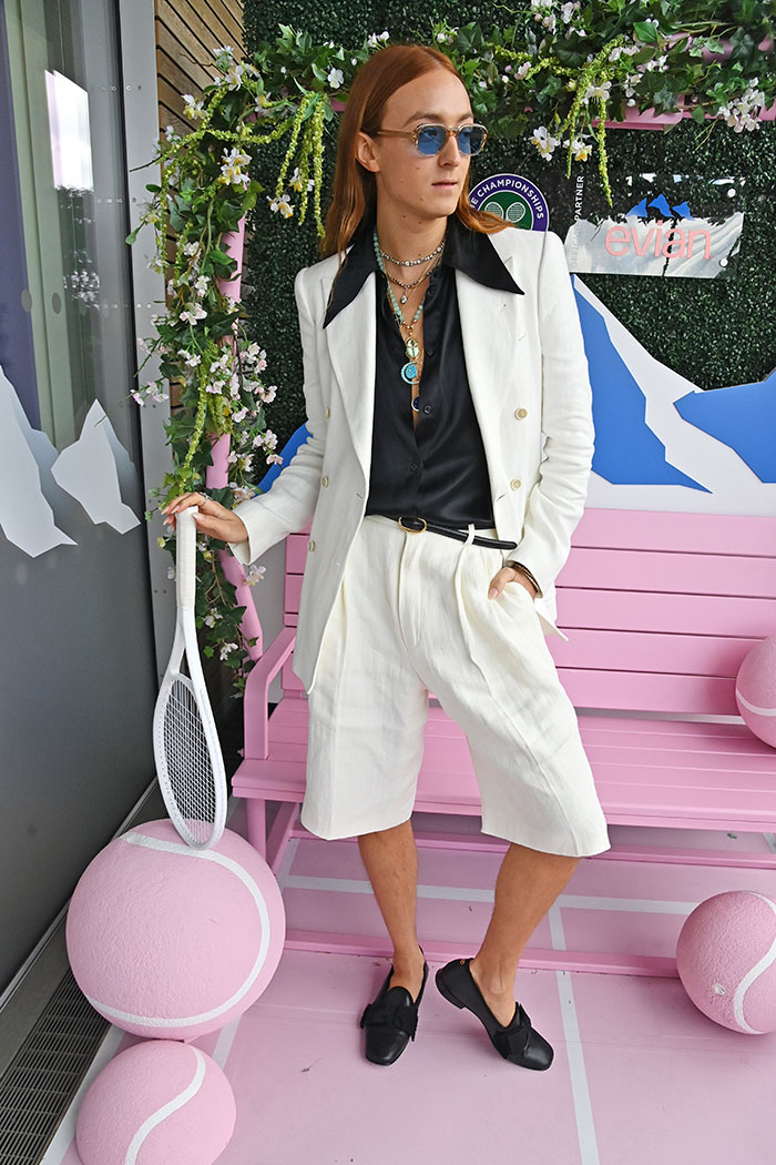 Fashionable celebrity at Wimbledon 2025 event wearing a white suit and holding a tennis racket by pink tennis balls.