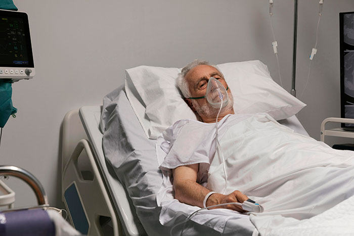 Elderly patient in hospital bed wearing oxygen mask with medical equipment nearby, showing hospital staff experiences