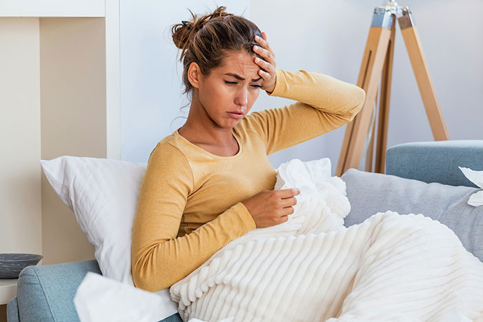 Woman sitting on a couch with a blanket, holding her head in discomfort, illustrating hospital staff witnessing common wild medical cases.