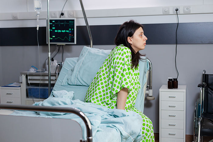 Patient in green hospital gown sitting on bed, with medical monitoring equipment and wheelchair in the background.