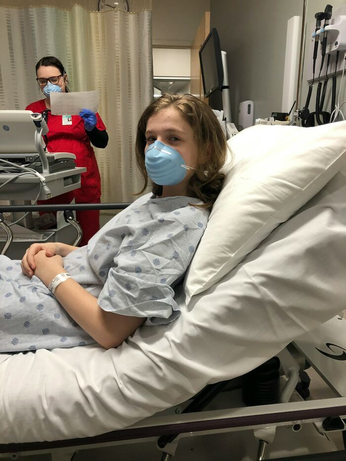 Young patient wearing a mask lies in a hospital bed with a nurse in protective gear checking documents nearby.