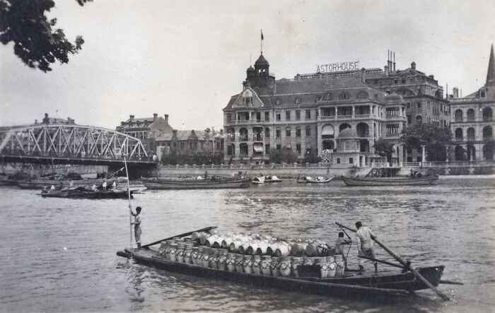 Black and white historical photograph showing river life and buildings from 100 years ago with boats and a distant bridge.