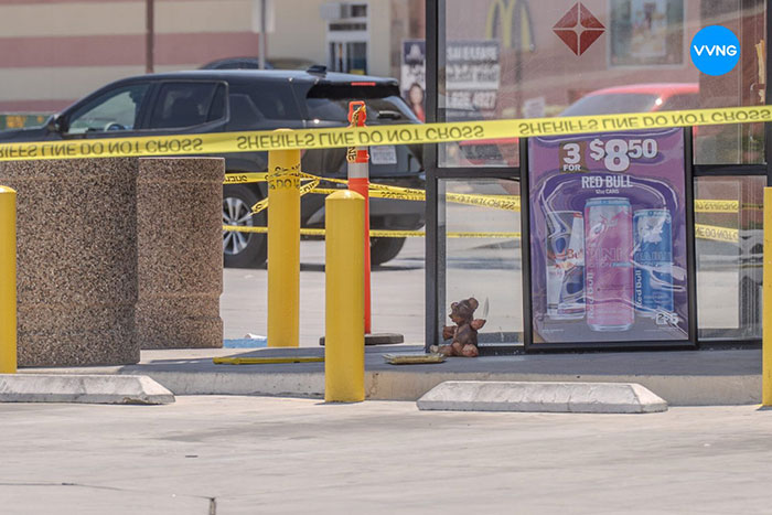 Teddy bear covered in apparent human remains sits outside convenience store behind yellow police tape near parking area.