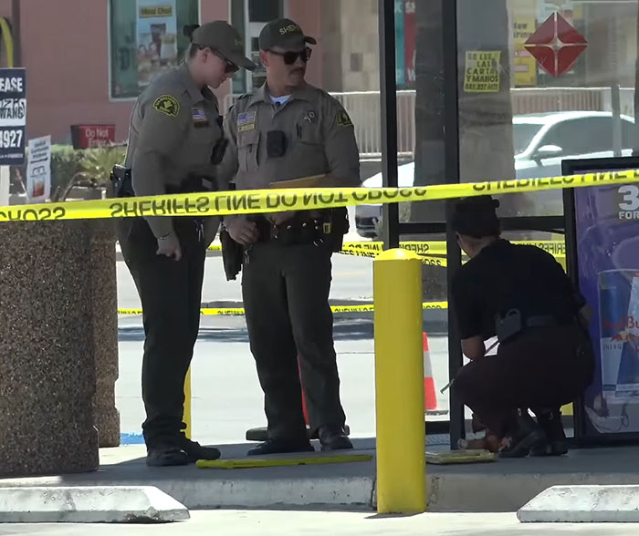Two sheriffs discussing at a convenience store crime scene with yellow police tape and an investigator crouching nearby.