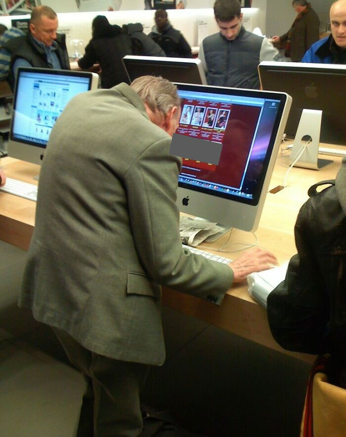 Older man focused on computer screen in a busy Apple store, showing a suspiciously unbothered attitude in public.