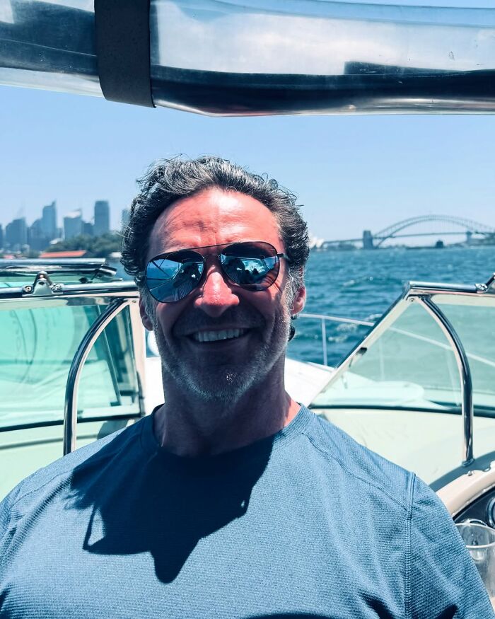 Hugh Jackman wearing sunglasses, smiling on a boat with water and city skyline in the background on a sunny day.