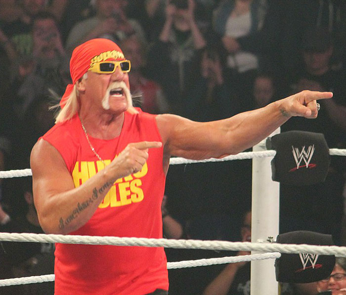 Hulk Hogan in wrestling ring wearing red bandana and shirt, pointing while engaging with the crowd at WWE event.