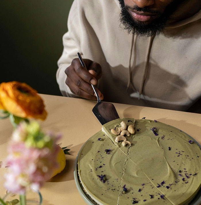 Man in beige hoodie cutting and serving a green cake slice topped with cashews on a beige table with flowers nearby.