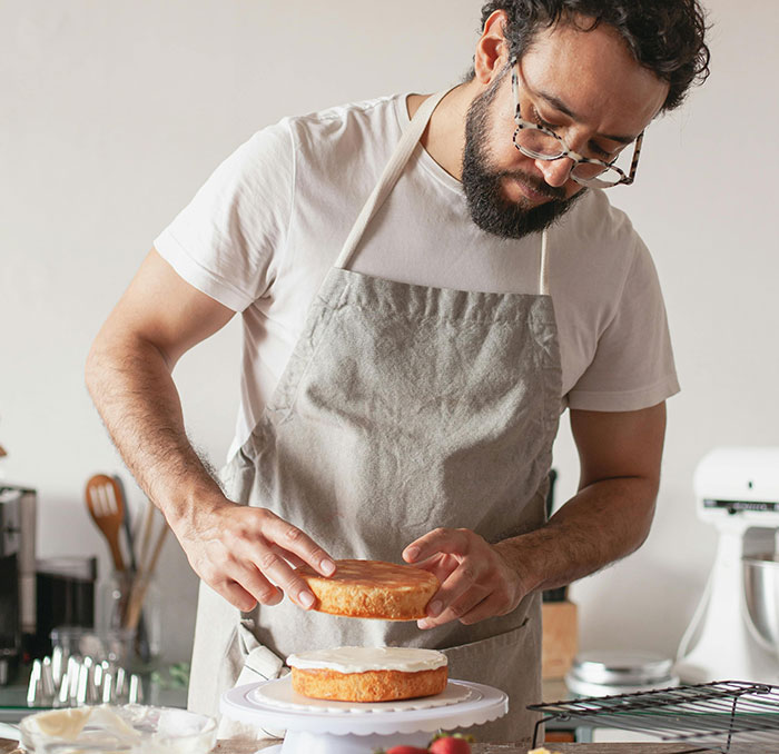 Man wearing glasses and apron carefully assembling a birthday cake in a kitchen, baking and preparing dessert.