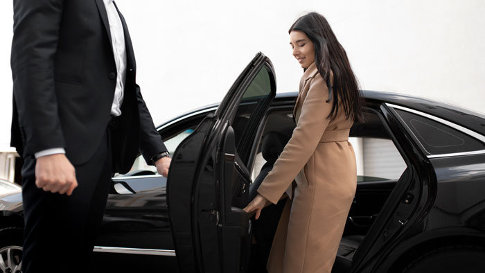 Woman in a tan coat entering a black car while a man in a suit holds the door, illustrating car service business discussion. - 9