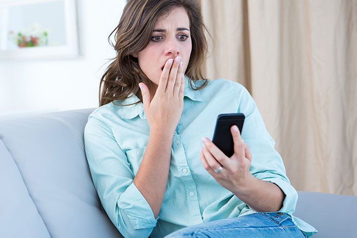 Woman sitting on couch, shocked and covering her mouth while looking at her smartphone, discovering husband texts mother.