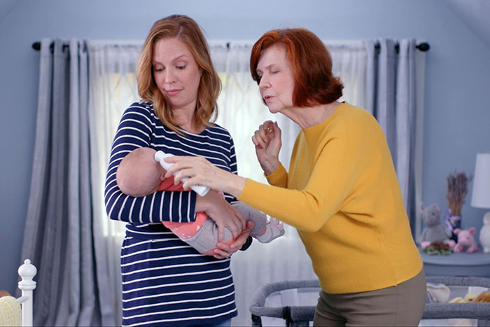 Woman holding baby in striped shirt while older woman in yellow sweater leans in closely, family tension evident in a nursery.