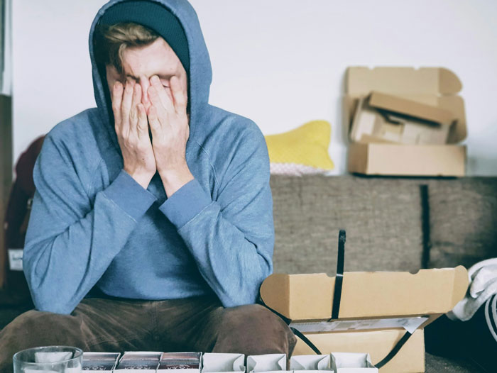 Man in hoodie covering face with hands, sitting amid unpacked boxes, illustrating travel difficulties and a weird quirk. - 15