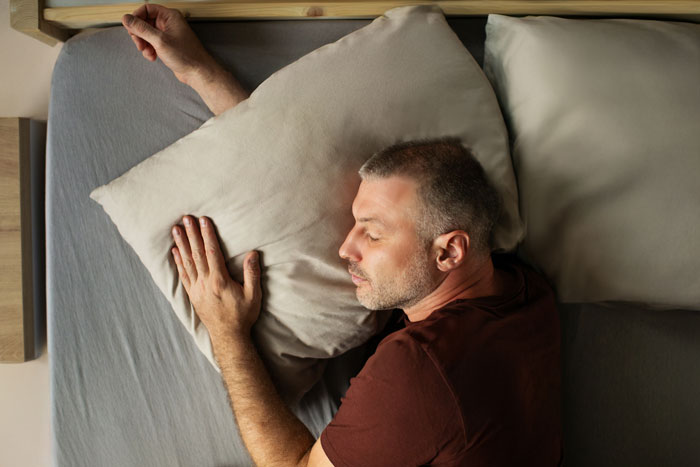 Man sleeping past noon in bed with pillow, highlighting parents wanting to wake him despite having pregnant wife at home.