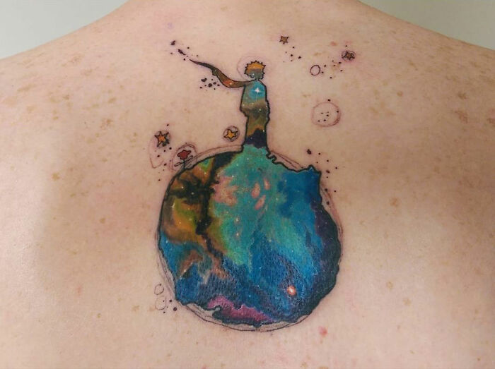 Colorful tattoo of a small planet with a figure standing on top surrounded by stars, one of the best tattoos trending now.