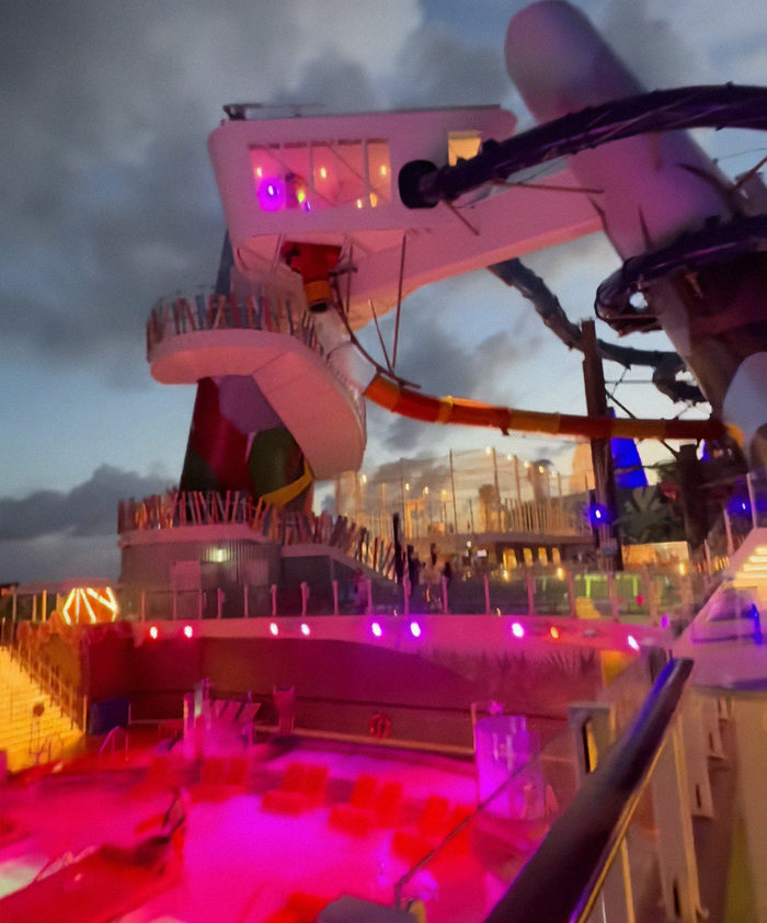 Cruise ship deck at dusk with vibrant lights near water slide and infinity pool edge area. - 2