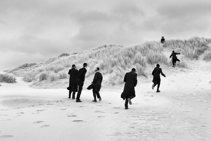Black and white street photo of six people dressed in dark coats walking and running on sandy dunes with cloudy sky.