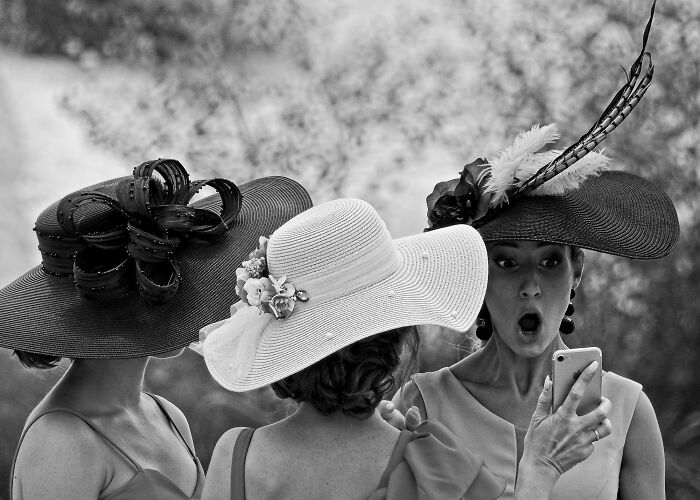 Three women wearing elaborate hats in a black and white street photo capturing a candid moment of surprise.