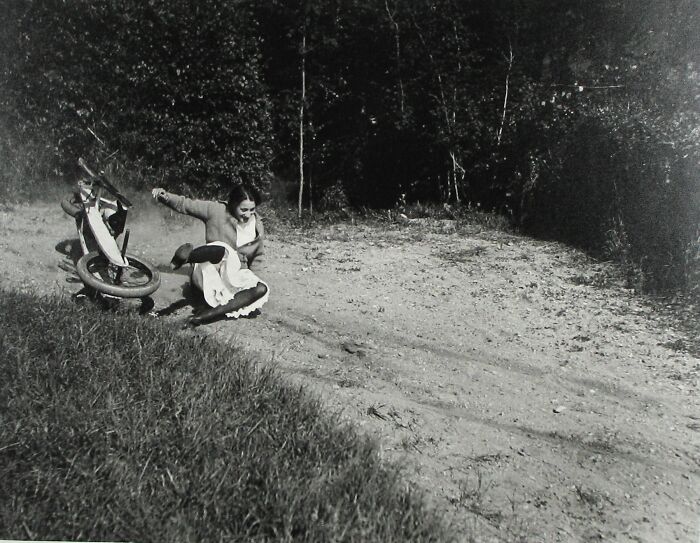Woman from the 1900s falling off a bicycle on a dirt path surrounded by trees, captured in an old photo.