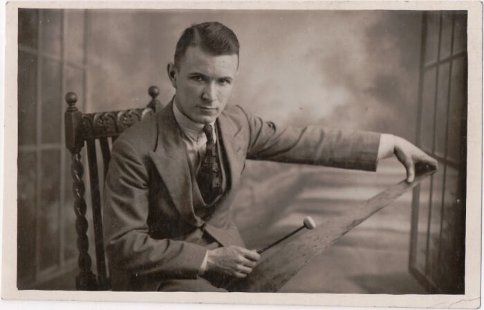 Vintage postcard showing a young man in 1930s attire holding a musical instrument in a classic sepia-toned setting.