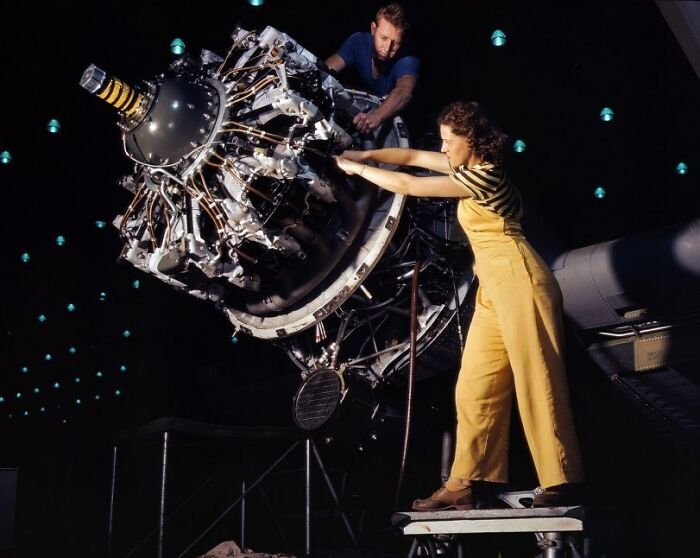 Woman in yellow overalls working on aircraft engine with man in dark shirt, showcasing women at work during WWII.